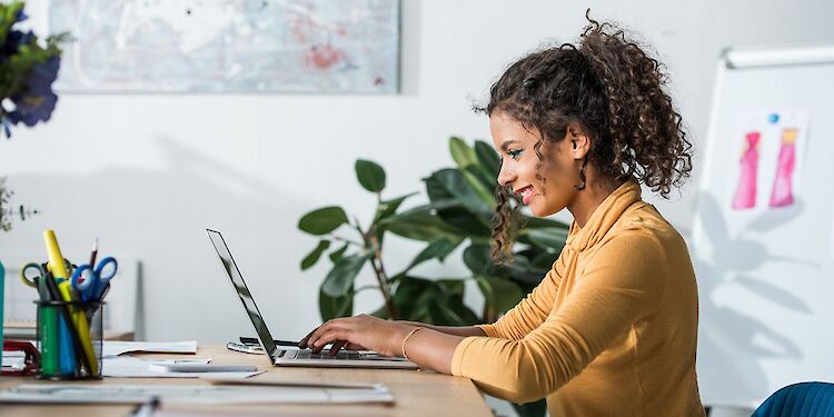 Vrouw aan bureau tikkend op laptop zittend in kantoor met op achtergrond een whiteboard en op bureaublad een pennenbakje