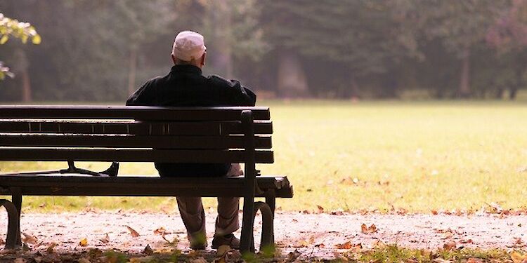 Achteraanzicht van een oudere man die op een bankje in het park zit, met een loopkruk naast hem