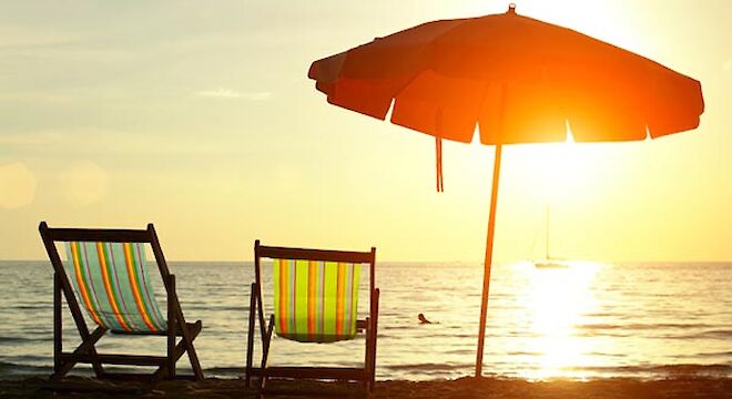 twee strandstoeltjes met een parasol ernaast met de zee en een zon die ondergaat op de achtergrond