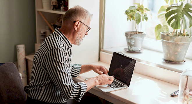 Man met bril zittend aan bureau met laptop voor hem. Op vensterbank staan planten.