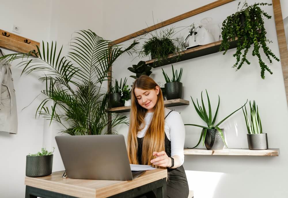 Vrouw zit aan tafel met kijkend naar laptop. Op achtergrond plankjes met planten